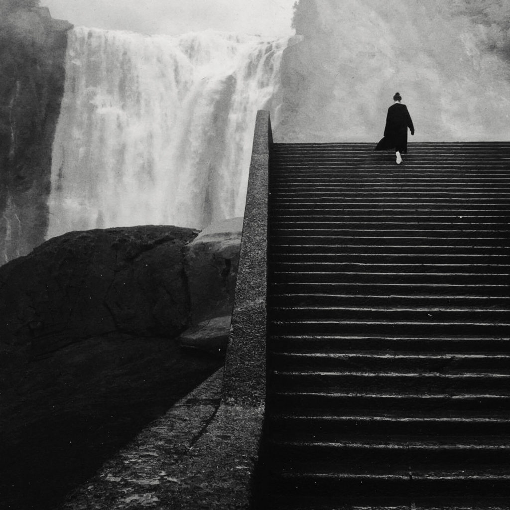 Aki walking up a tall outdoor staircase, with a waterfall off in the distance